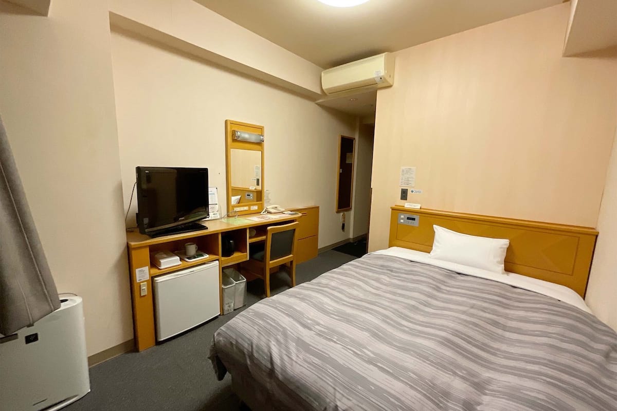 Ein Hotelzimmer mit Bett, Schreibtisch mit Fernseher, kleinem Kühlschrank, Spiegel und Klimaanlage.