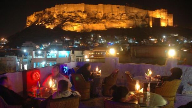 A rooftop dining area with tables and chairs, a large illuminated building in the background, and a view of the cityscape.