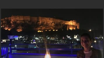 A person seated at a table with a lit lamp, overlooking a large illuminated cliff at night.