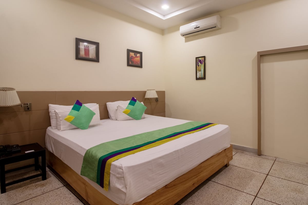 A hotel room with a wooden bed, white bedding, a bedside table with a lamp, and a wall-mounted air conditioner.