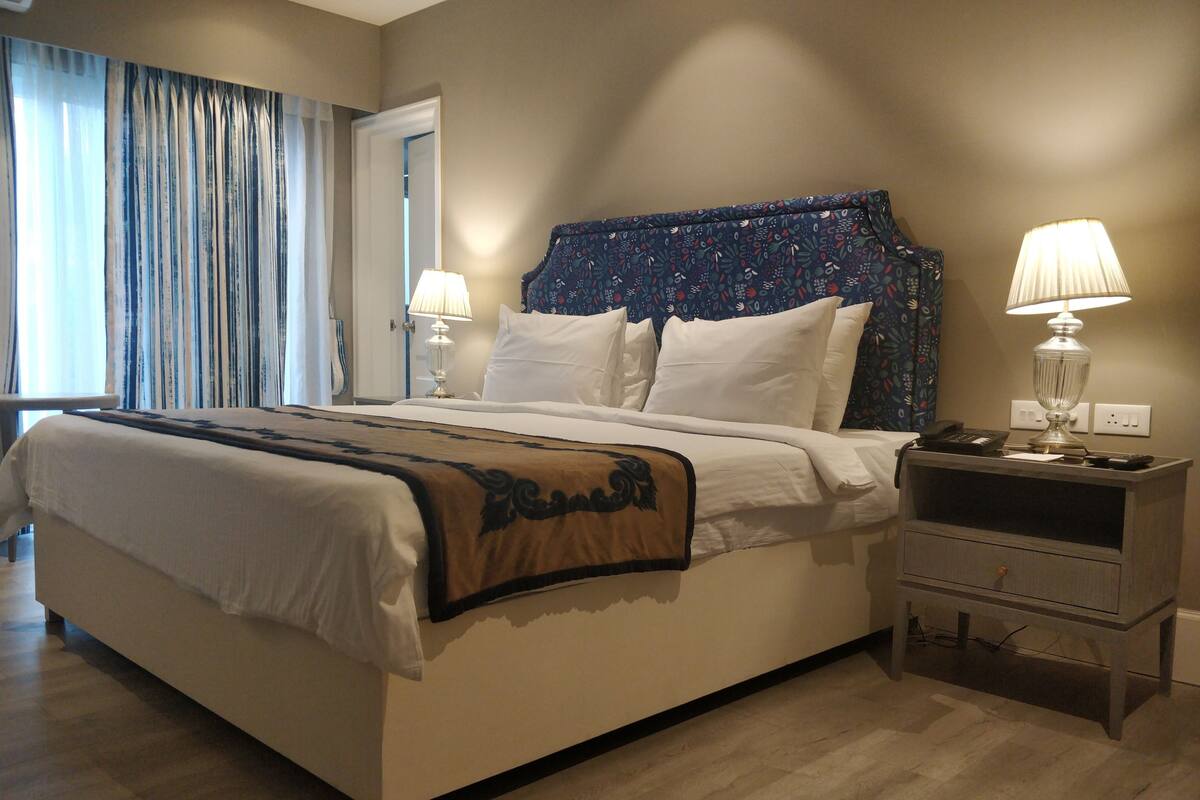 A neatly made bed with a patterned headboard, bedside lamps, and a bedside table with a phone and remote controls.