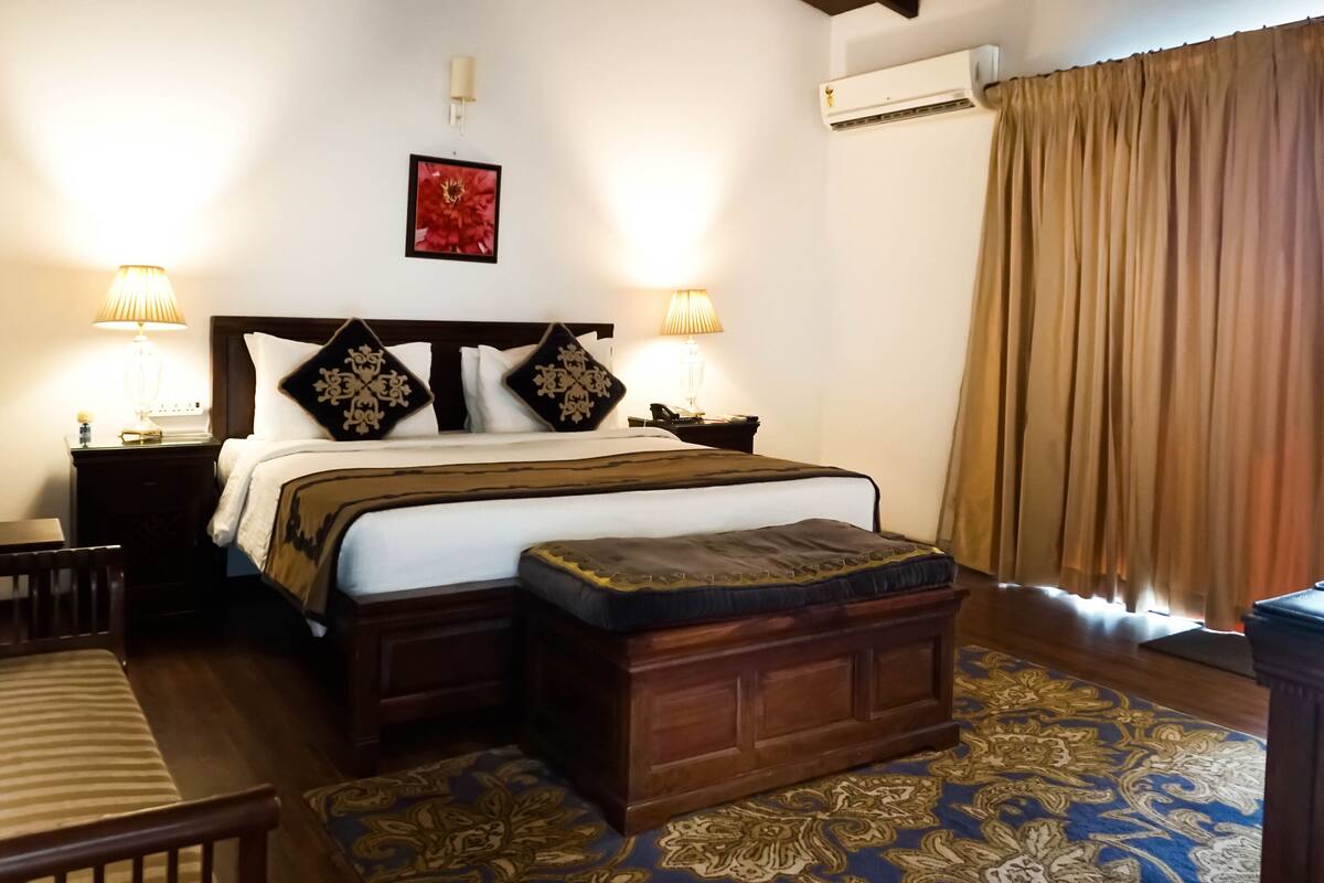 A hotel room with a large bed, bedside lamps, a nightstand, a wooden bench, a patterned rug, and a window with curtains.