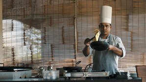 A chef in a kitchen with a bamboo blind background, cooking utensils, and a stove.