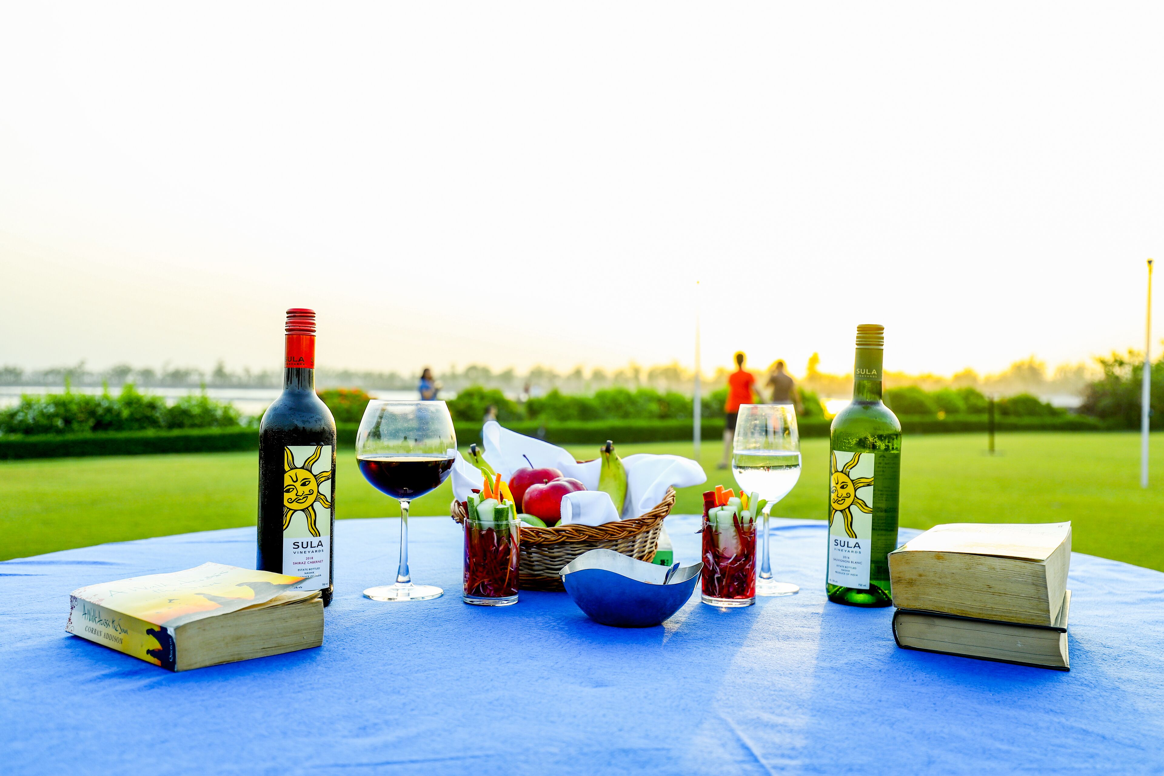 A table set outdoors with a bottle of Sula wine, two glasses of wine, a basket of fruits, and two books.