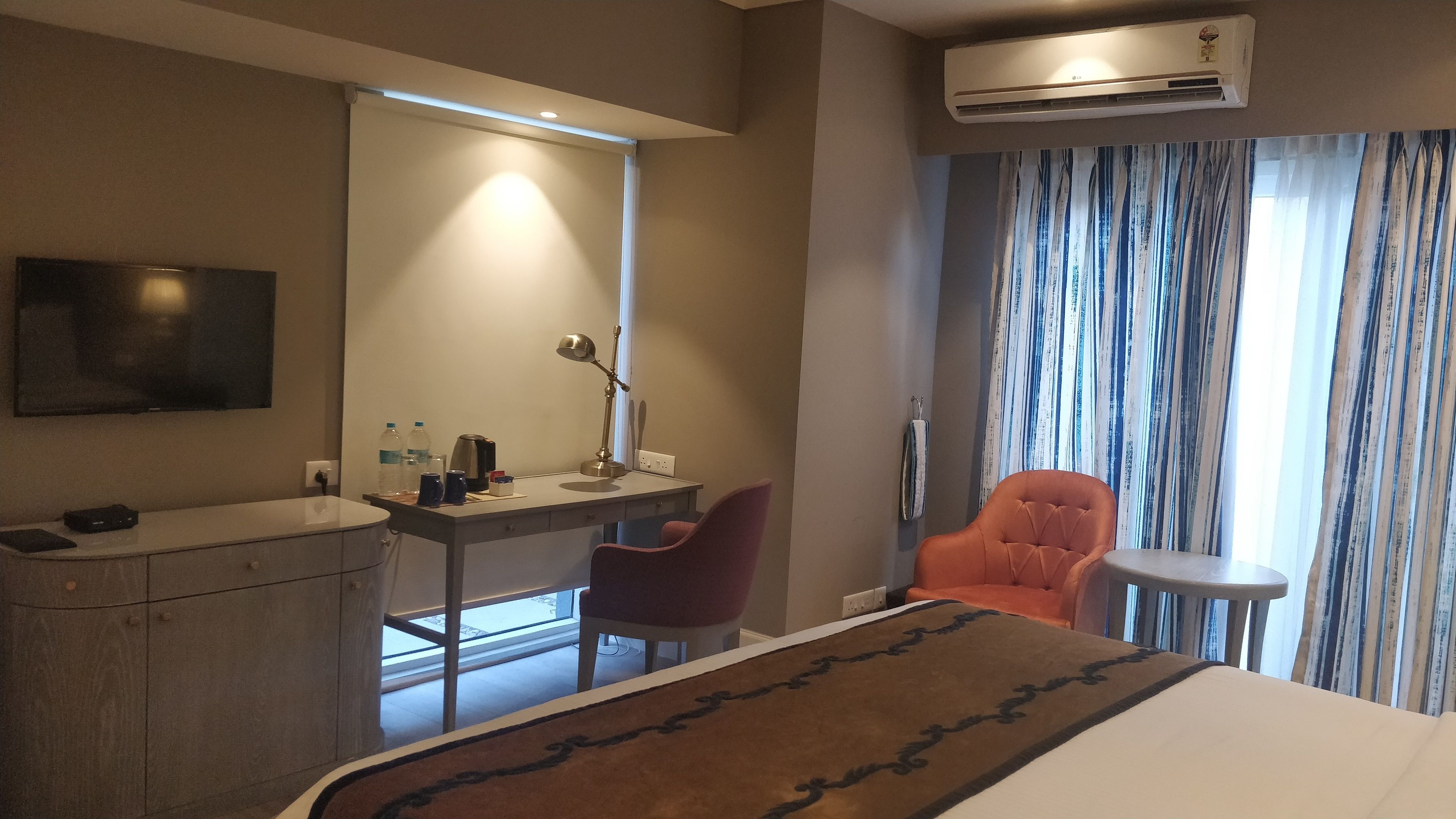 Habitación de hotel con cama, escritorio, silla, televisor, aire acondicionado y ventana con cortinas.