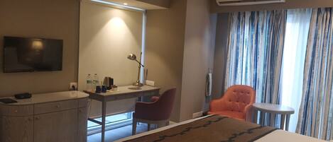 Habitación de hotel con cama, escritorio, silla, televisor, aire acondicionado y ventana con cortinas.