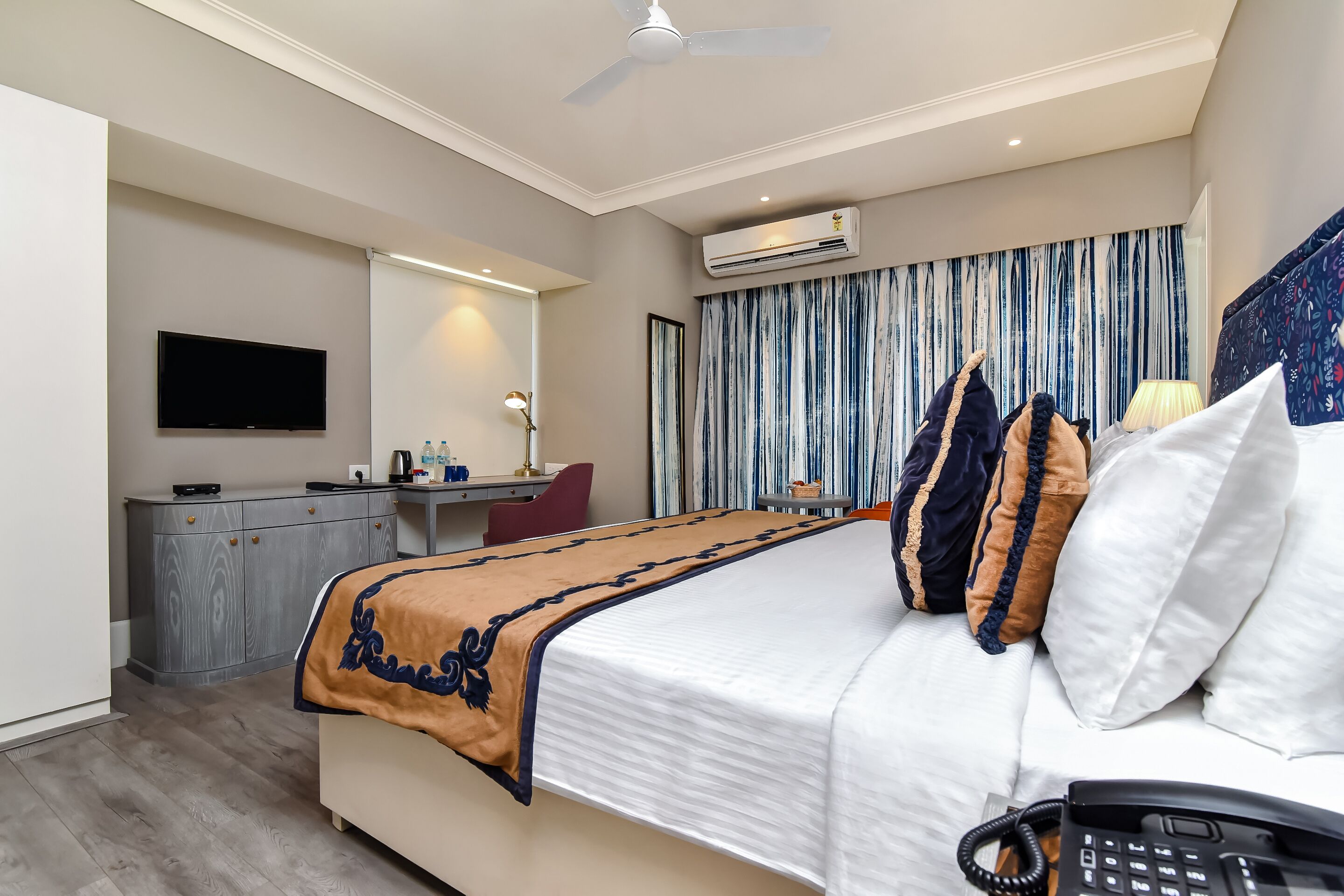 A hotel room with a bed, a desk, a flat-screen TV, an air conditioner, and a ceiling fan.