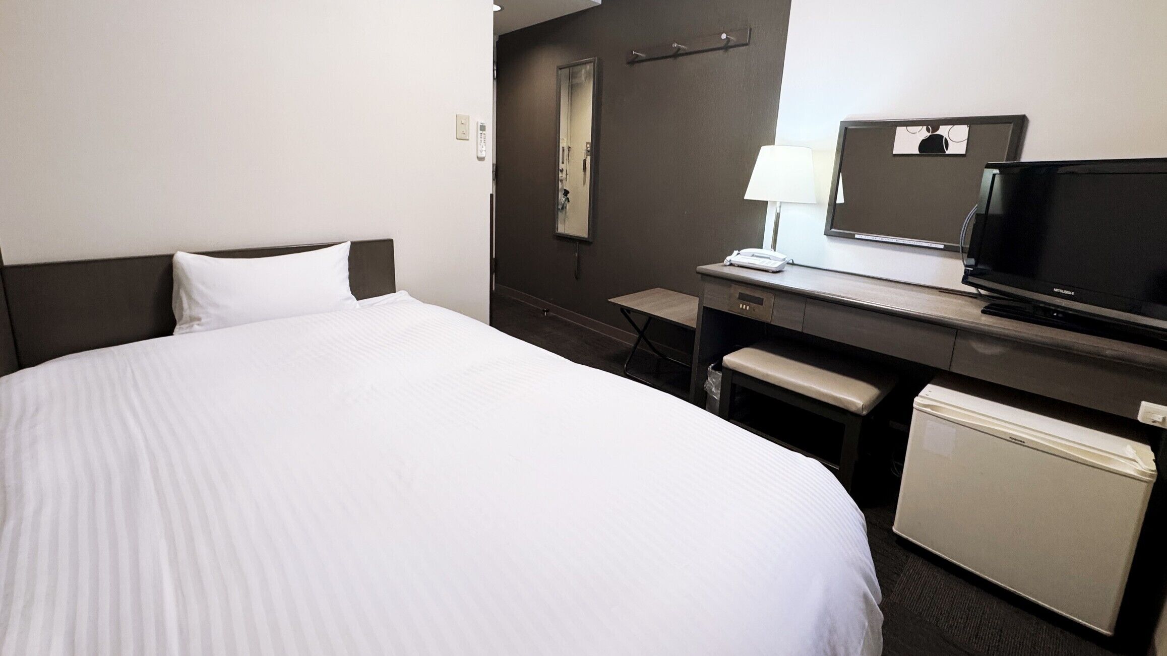 A hotel room with a bed, a desk with a television, a mirror, and a small refrigerator.