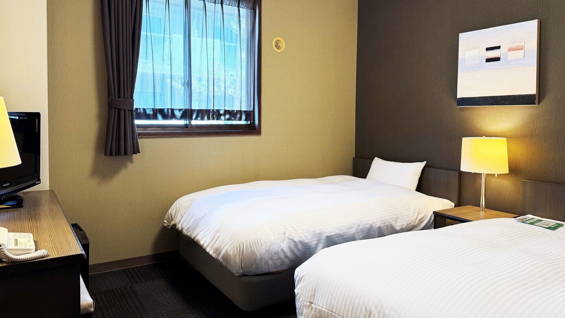 A hotel room with two beds, a desk with a telephone, a window with curtains, a wall-mounted light switch, and a framed picture.