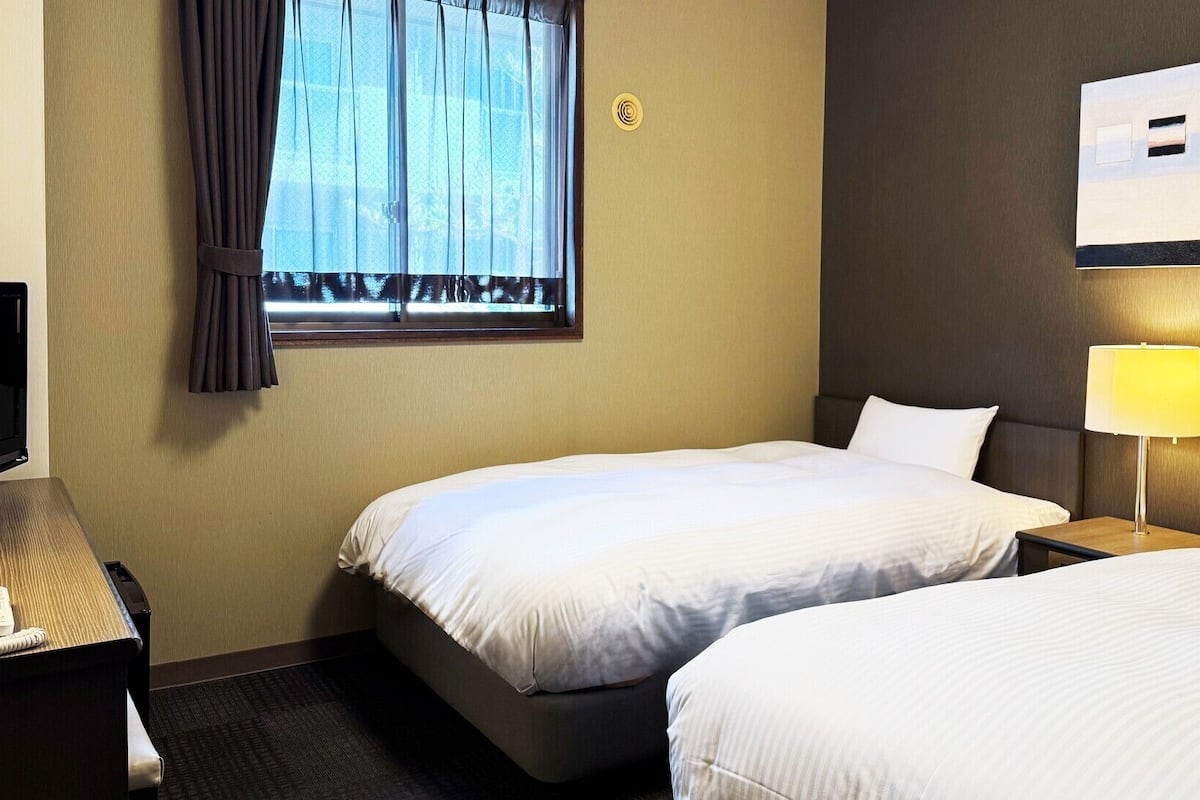 Une chambre d’hôtel avec deux lits, un bureau avec un téléphone, une fenêtre avec des rideaux, un interrupteur fixé au mur et un tableau encadré.