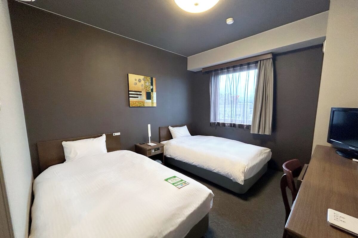 Ein Hotelzimmer mit zwei Betten, einem Schreibtisch mit Stuhl, einem Fernseher und einem Fenster mit Vorhängen.