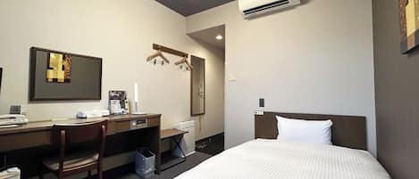 A hotel room with a bed, a desk, a chair, a trash bin, and an air conditioning unit.
