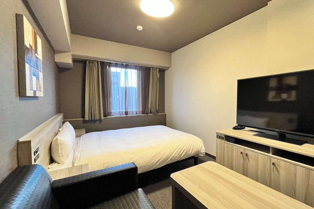 A hotel room with a bed, a TV, a small seating area, and a window with curtains.