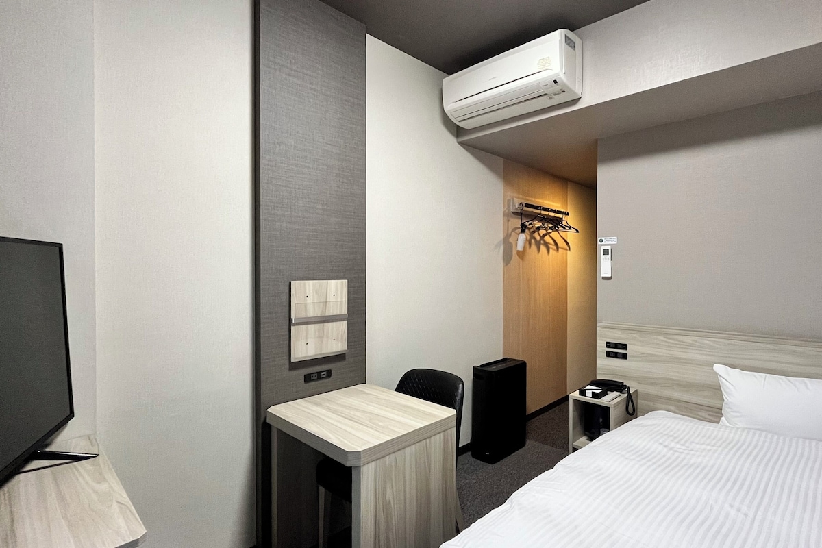 A hotel room with a bed, a desk, a chair, an air conditioning unit, a speaker, and a coat rack.