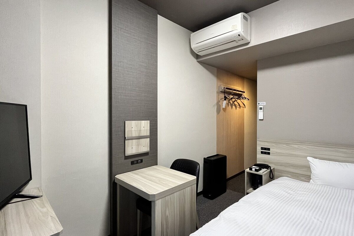 A hotel room with a bed, a desk with a chair, a television, an air conditioning unit, and a wall-mounted light fixture.