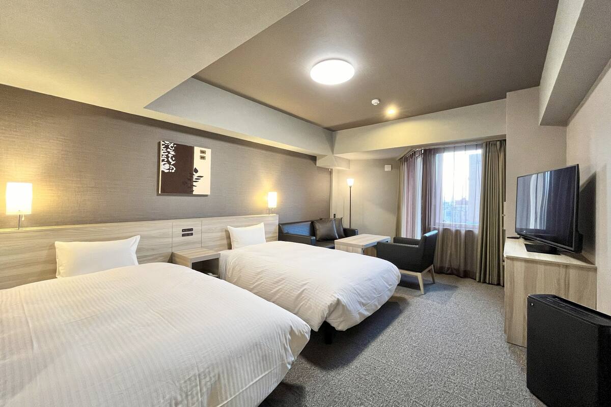 Ein Hotelzimmer mit zwei Betten, einem Schreibtisch, einem Stuhl, einem Fernseher und einem Fenster mit Vorhängen.