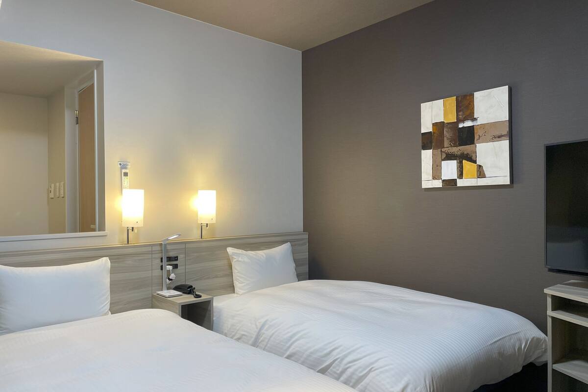 Ein Hotelzimmer mit zwei Betten, einem Nachttisch mit Lampe, einem Wand-Fernseher und einem Gemälde.