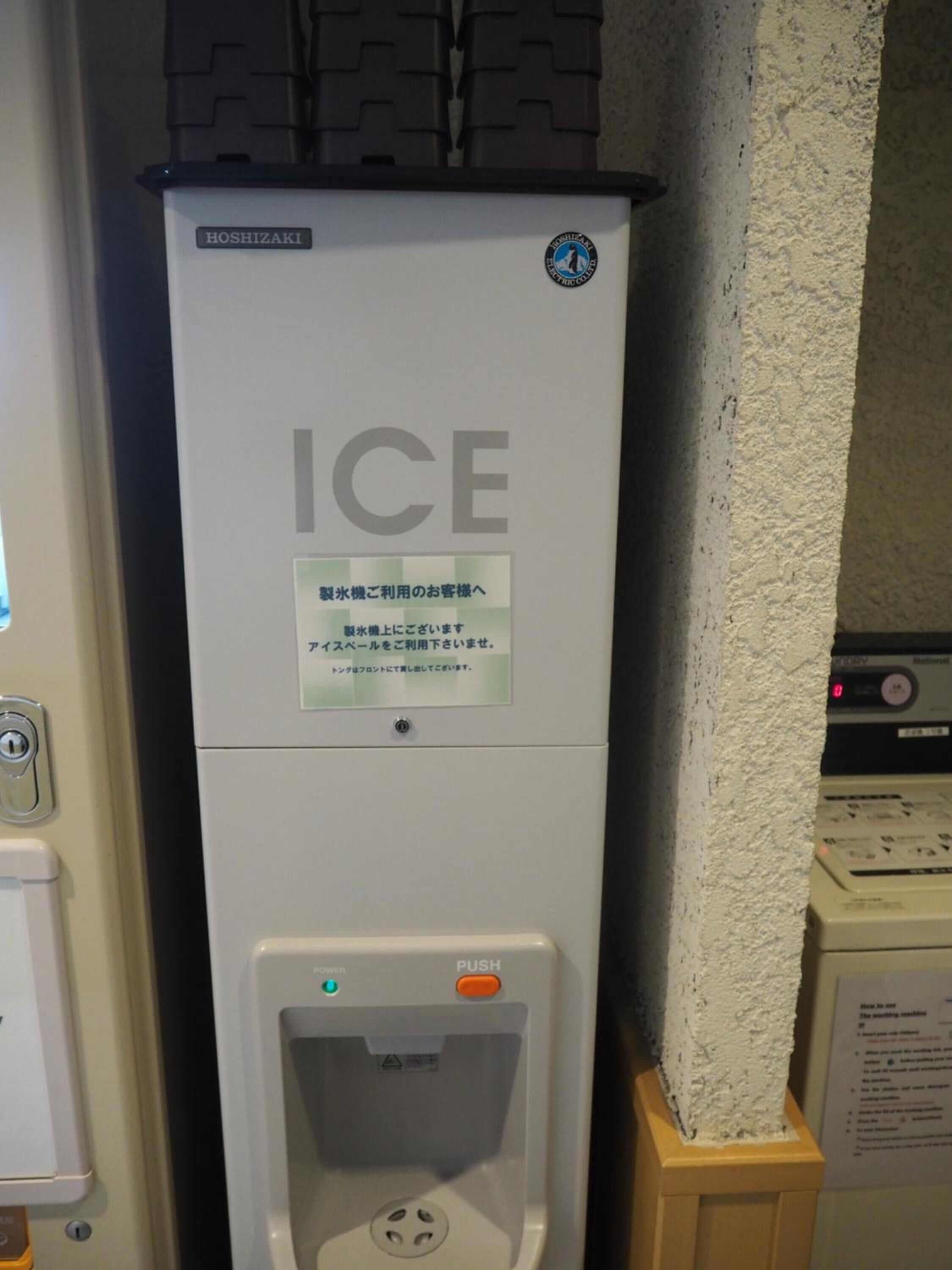 An ice dispenser with instructions in Japanese, located in a room with a textured wall and a control panel in the background.