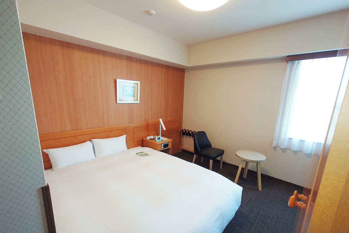 A hotel room with a large bed, a small table, a chair, and a window with curtains.