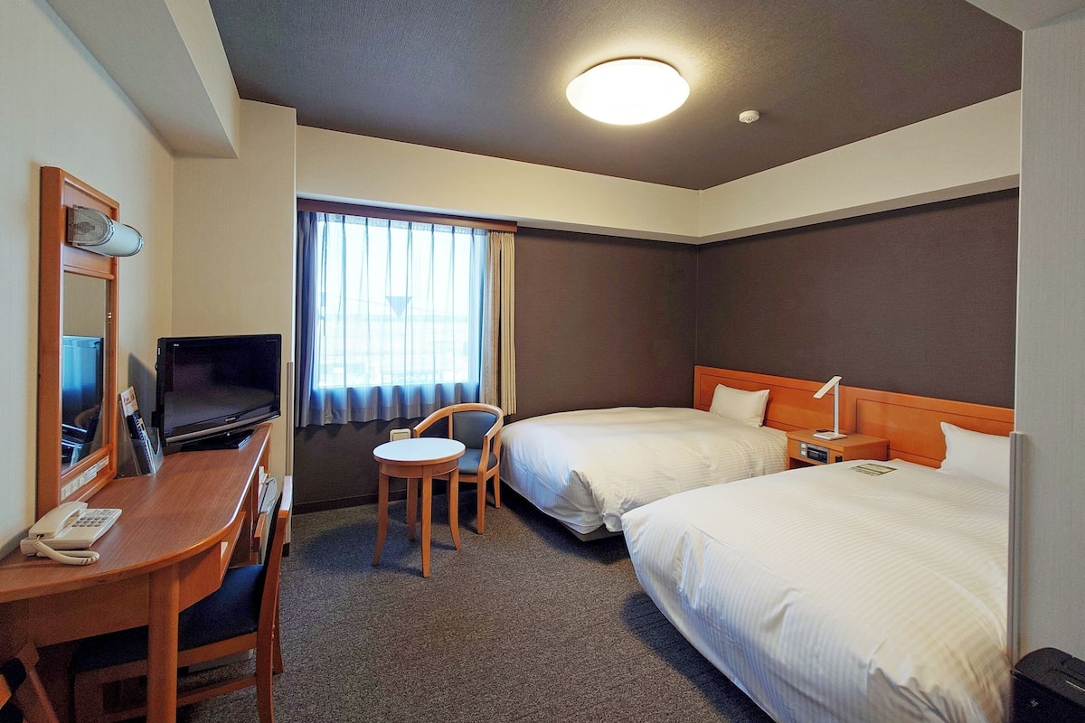 A hotel room with a desk, two beds, a TV, a chair, and a window with curtains.