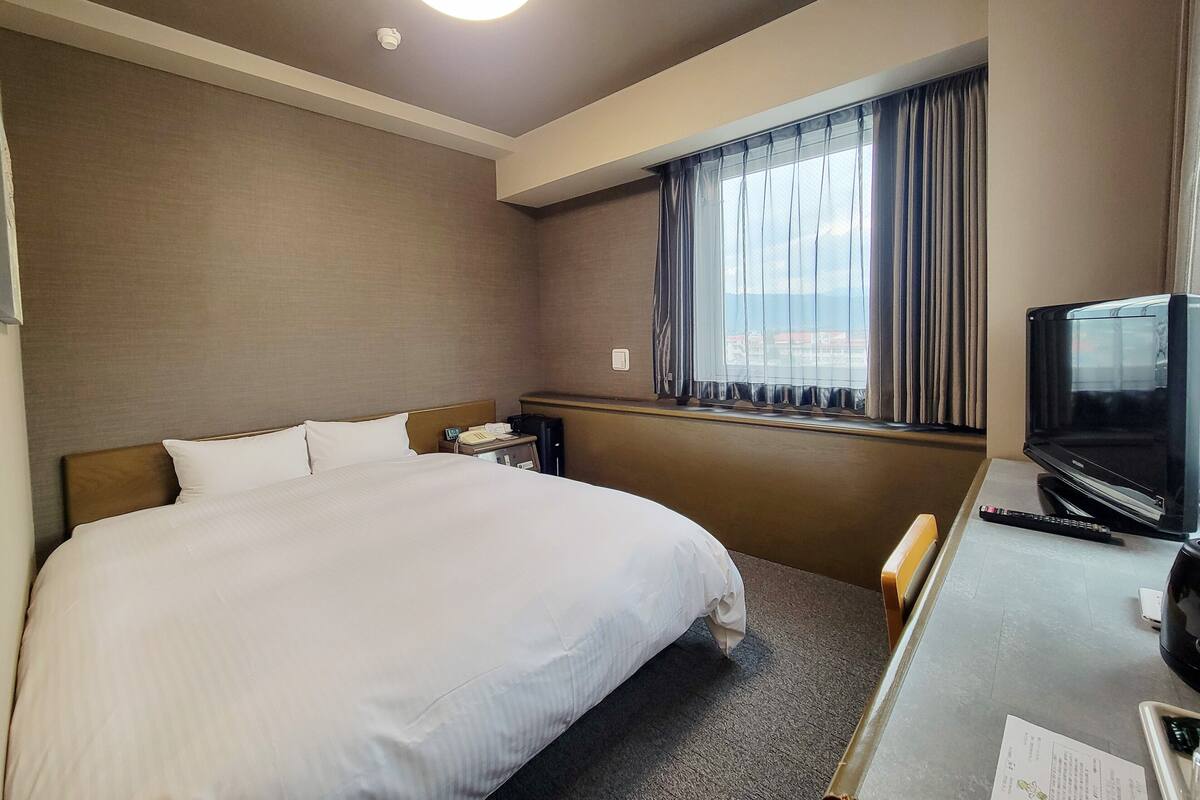 A hotel room with a large bed, a desk with a television, a window with curtains, and a view of the outside.