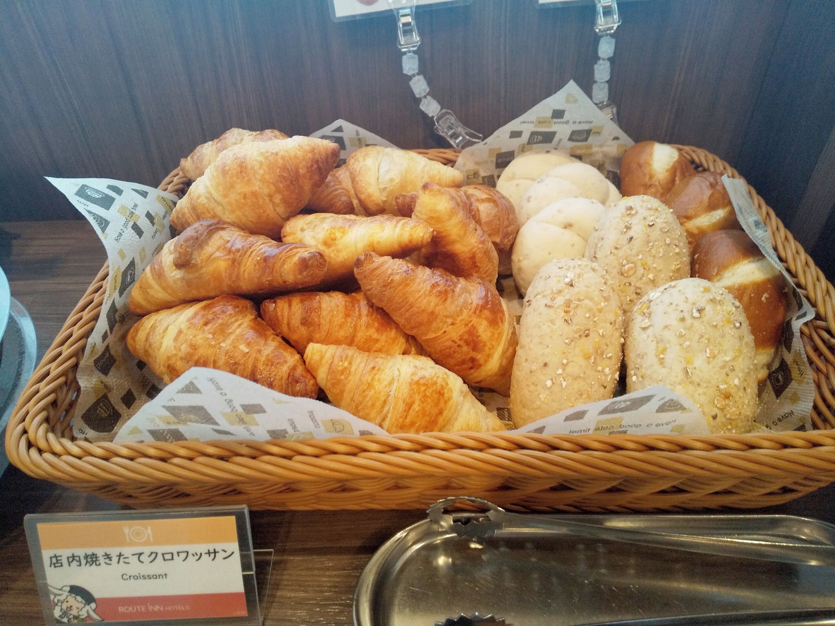 A basket of assorted baked goods including croissants and bread, placed on a table with a sign indicating the items.