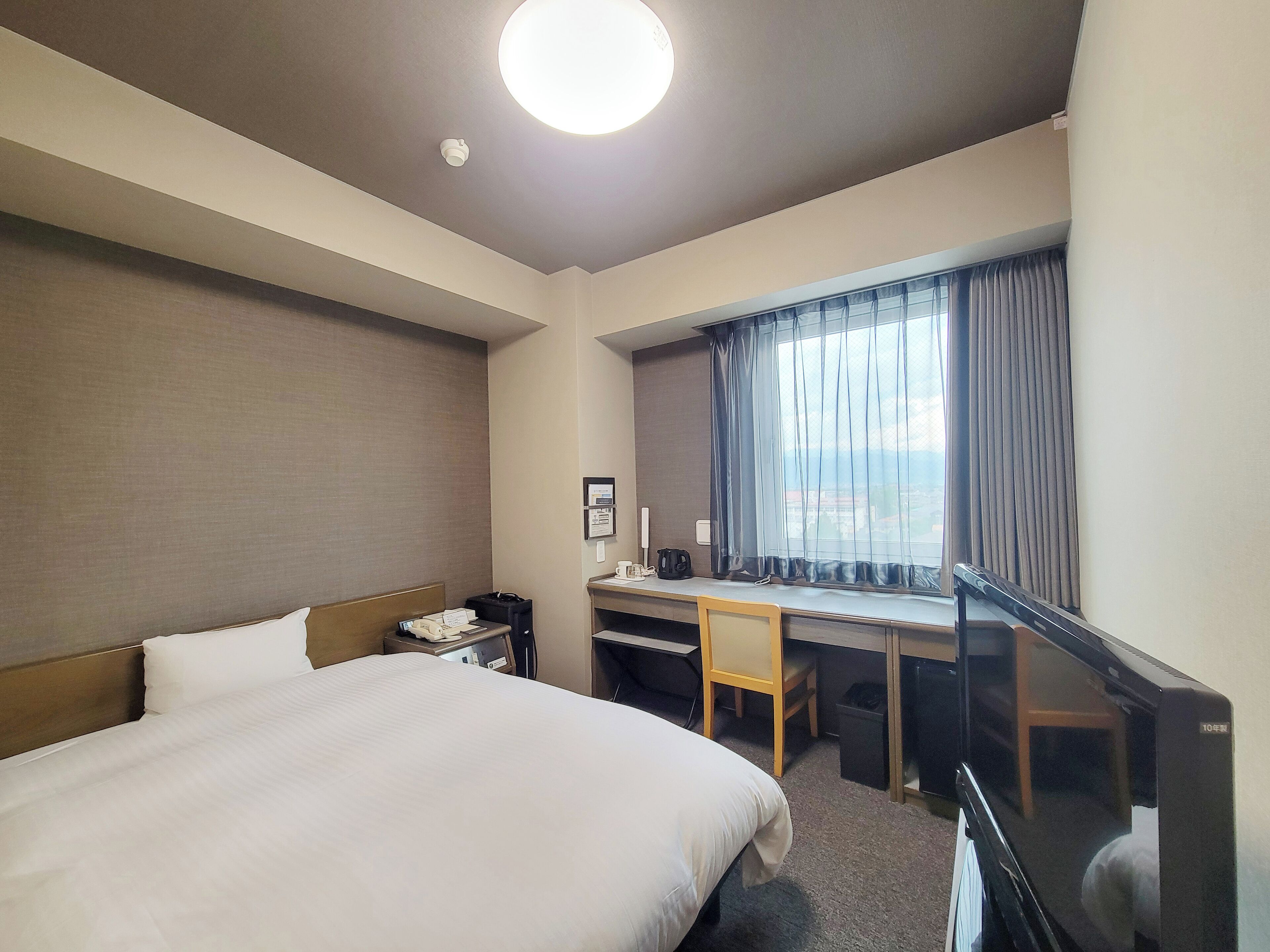 Hotellihuone, jossa on sänky, työpöytä, tuoli ja ikkunasta avautuva näkymä kaupunkiin.