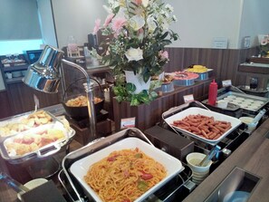 Un bufé con diversos alimentos, como pasta, perros calientes y pasteles, junto con un arreglo floral decorativo.