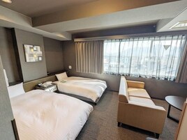 Habitación de hotel con dos camas, un sofá, una mesita y vistas a la ciudad a través de amplios ventanales.