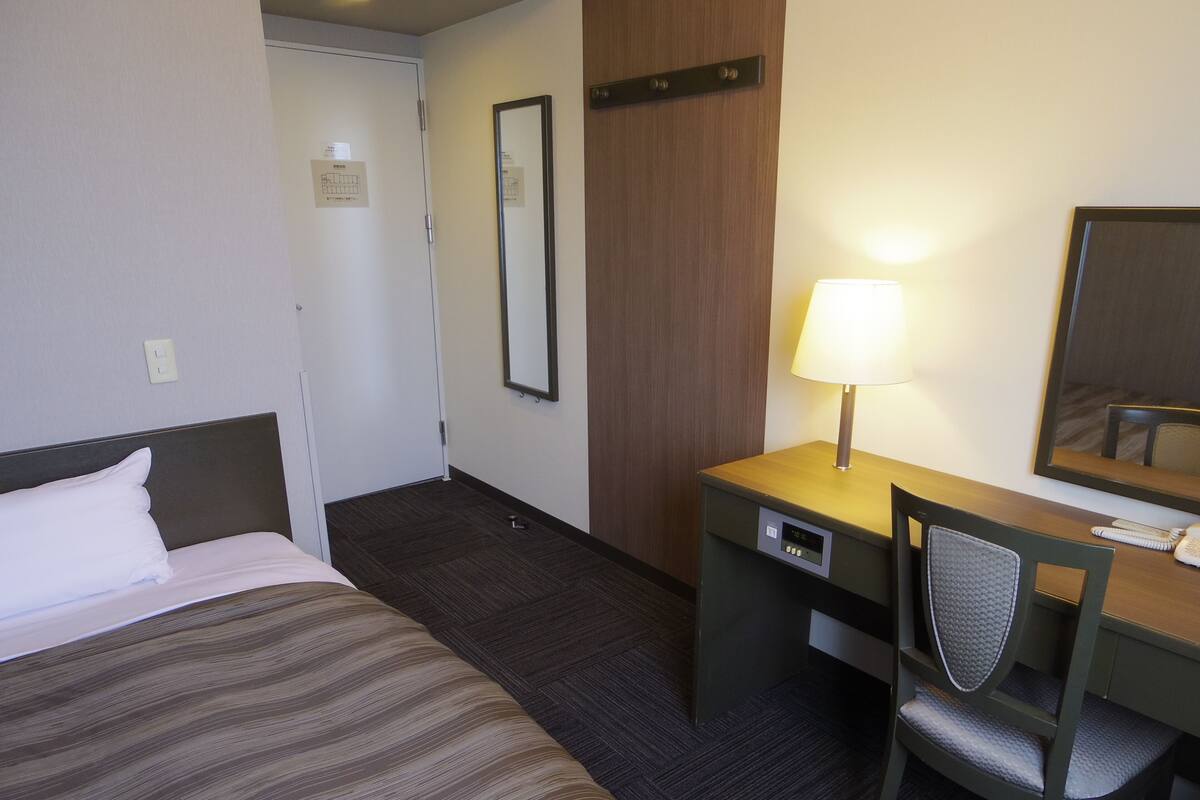 A hotel room with a bed, a desk, a chair, a lamp, and a mirror.