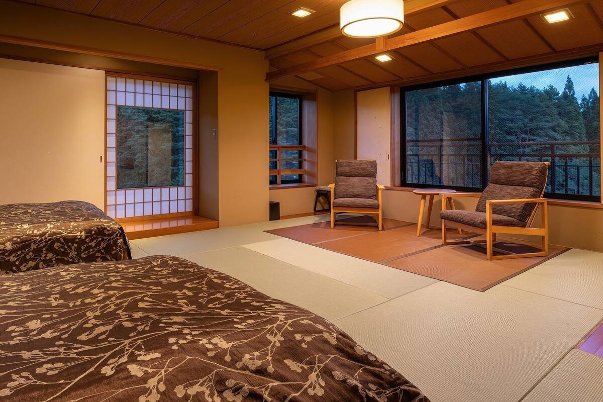 A room with two beds, a small table, two chairs, and large windows with a view of trees.