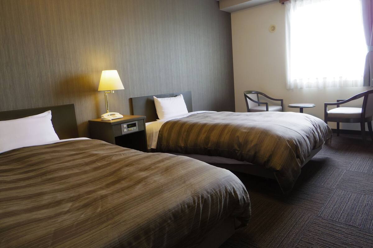 A hotel room with two beds, a bedside table with a lamp, a chair, and a small table.