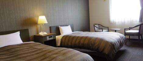 A hotel room with two beds, a bedside table with a lamp, a chair, and a small table.