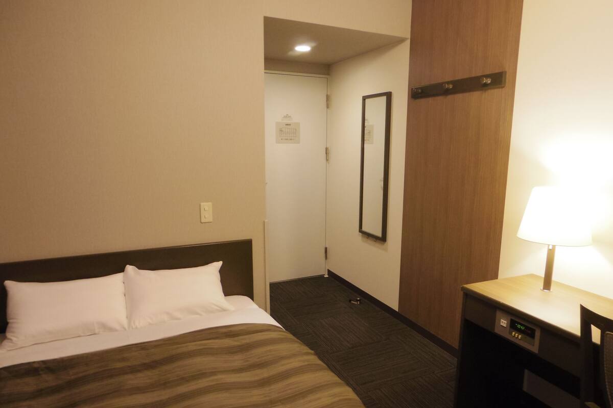 A hotel room with a bed, a desk, a chair, a mirror, and a lamp.