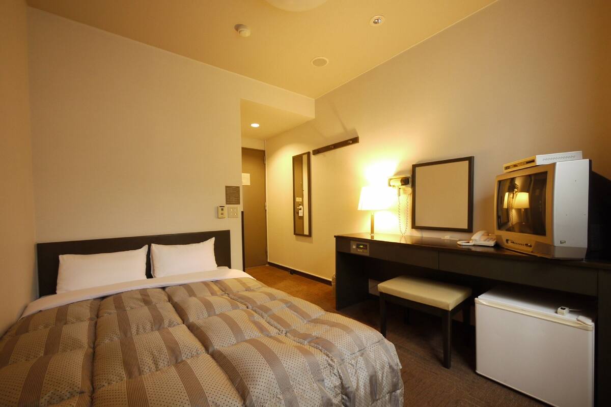 A hotel room with a bed, a desk, a small refrigerator, a lamp, and a television.