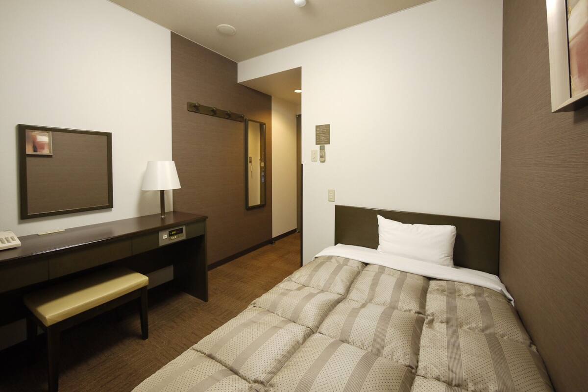 A hotel room with a bed, a desk, a chair, a mirror, and a wall-mounted phone.