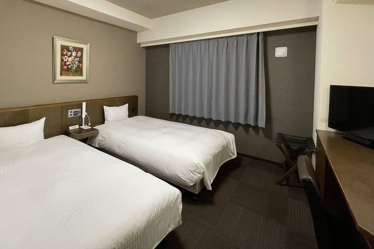 A hotel room with two beds, a desk, a chair, a television, a painting on the wall, and a window with curtains.