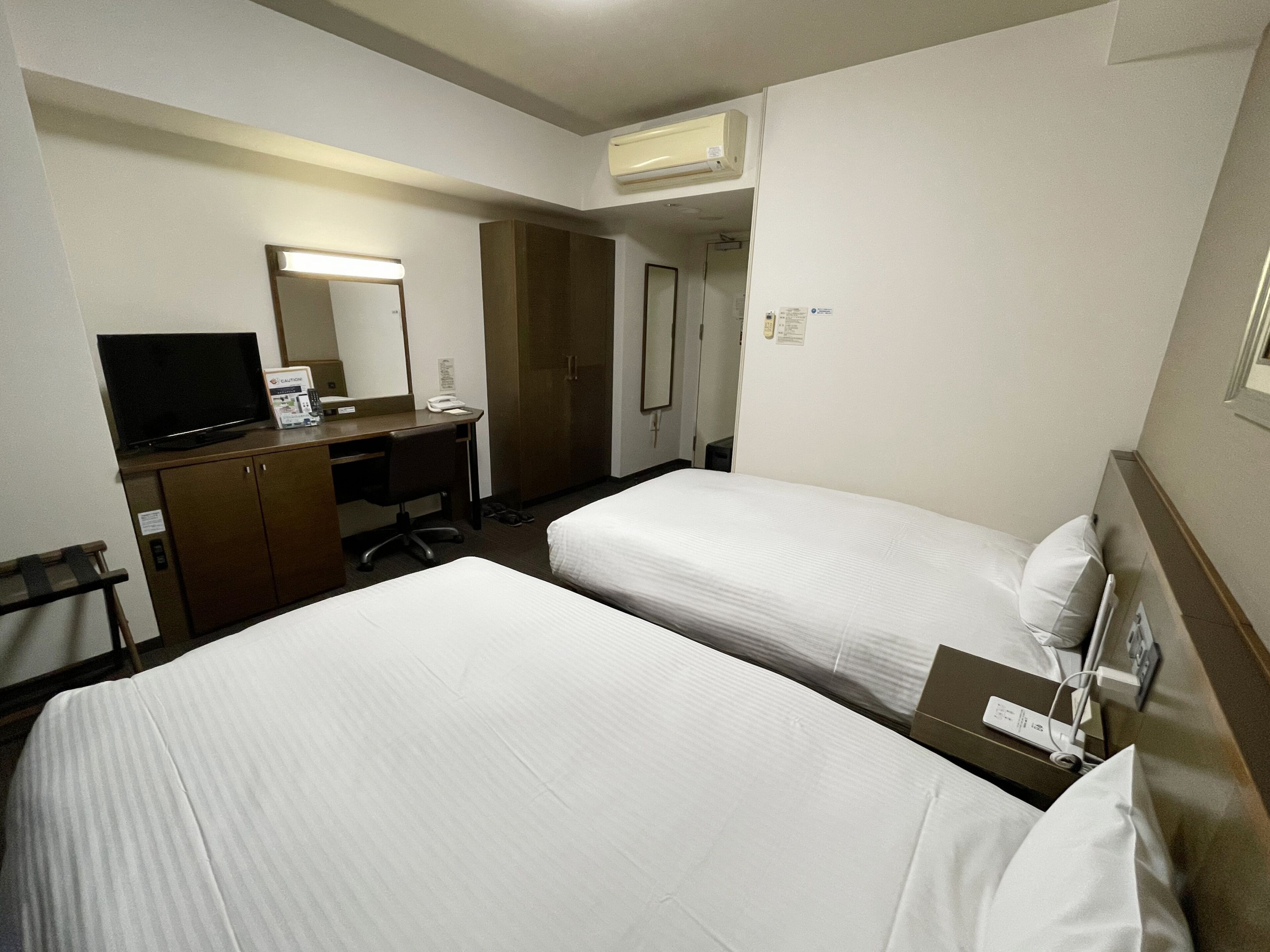 Habitación de hotel con dos camas, un escritorio con televisor, un espejo y un equipo de aire acondicionado.