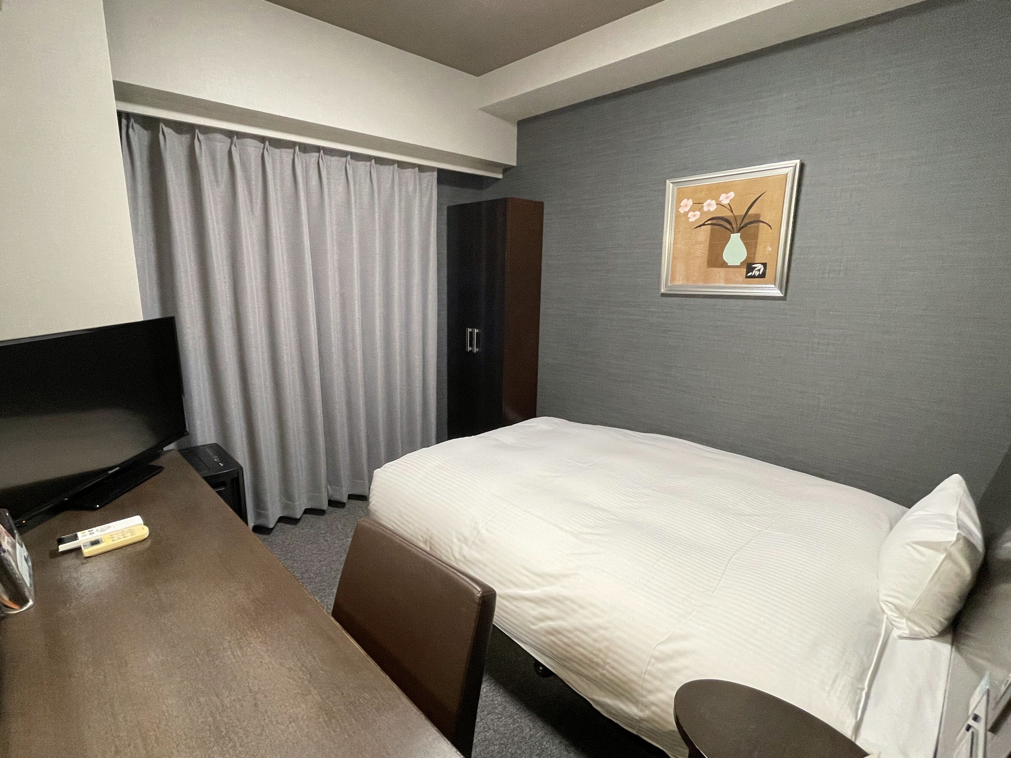 A hotel room with a bed, a desk, a chair, a TV, a wardrobe, and a framed picture on the wall.