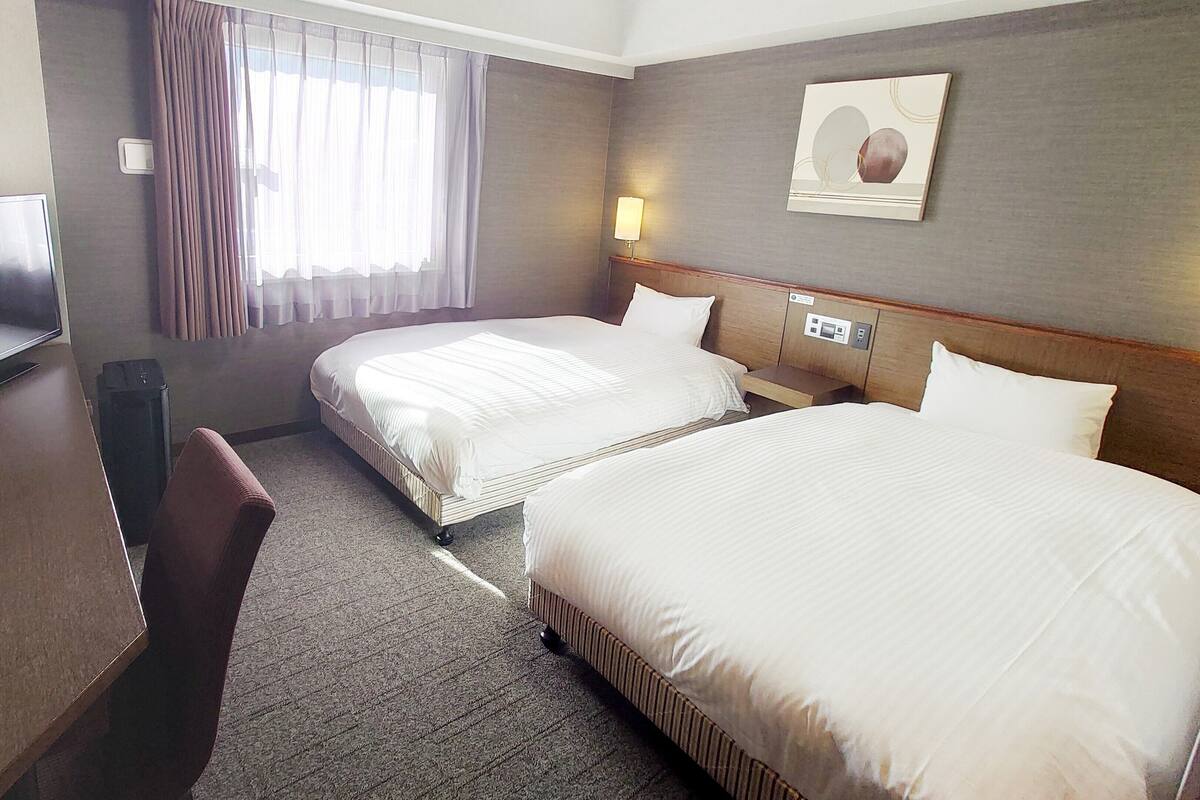 A hotel room with two beds, a desk, a chair, a TV, and a window with curtains.