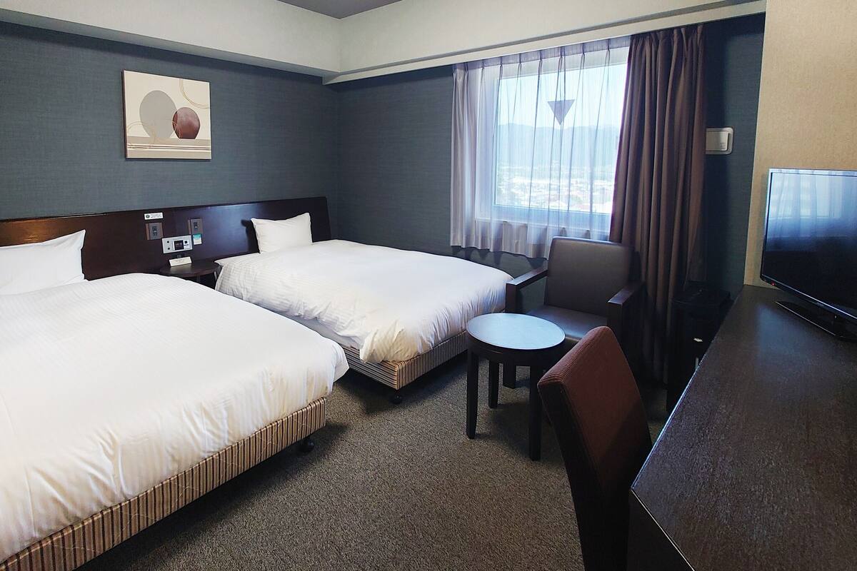 A hotel room with two beds, a small table, a chair, a TV, and a painting on the wall.