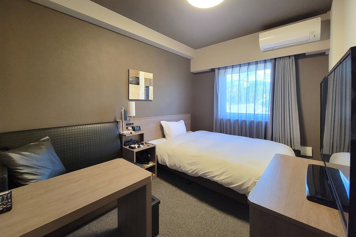 Quarto de hotel com cama, escrivaninha, cadeira, mesa de cabeceira, ar condicionado e janela com cortinas.