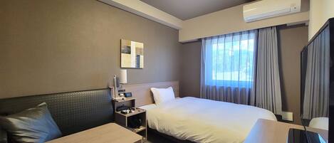 A hotel room with a bed, a desk, a chair, a nightstand, an air conditioner, and a window with curtains.