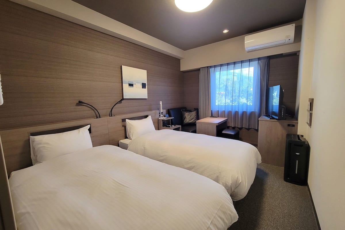 Quarto de hotel com duas camas, uma mesa, uma cadeira, televisão e ar condicionado.