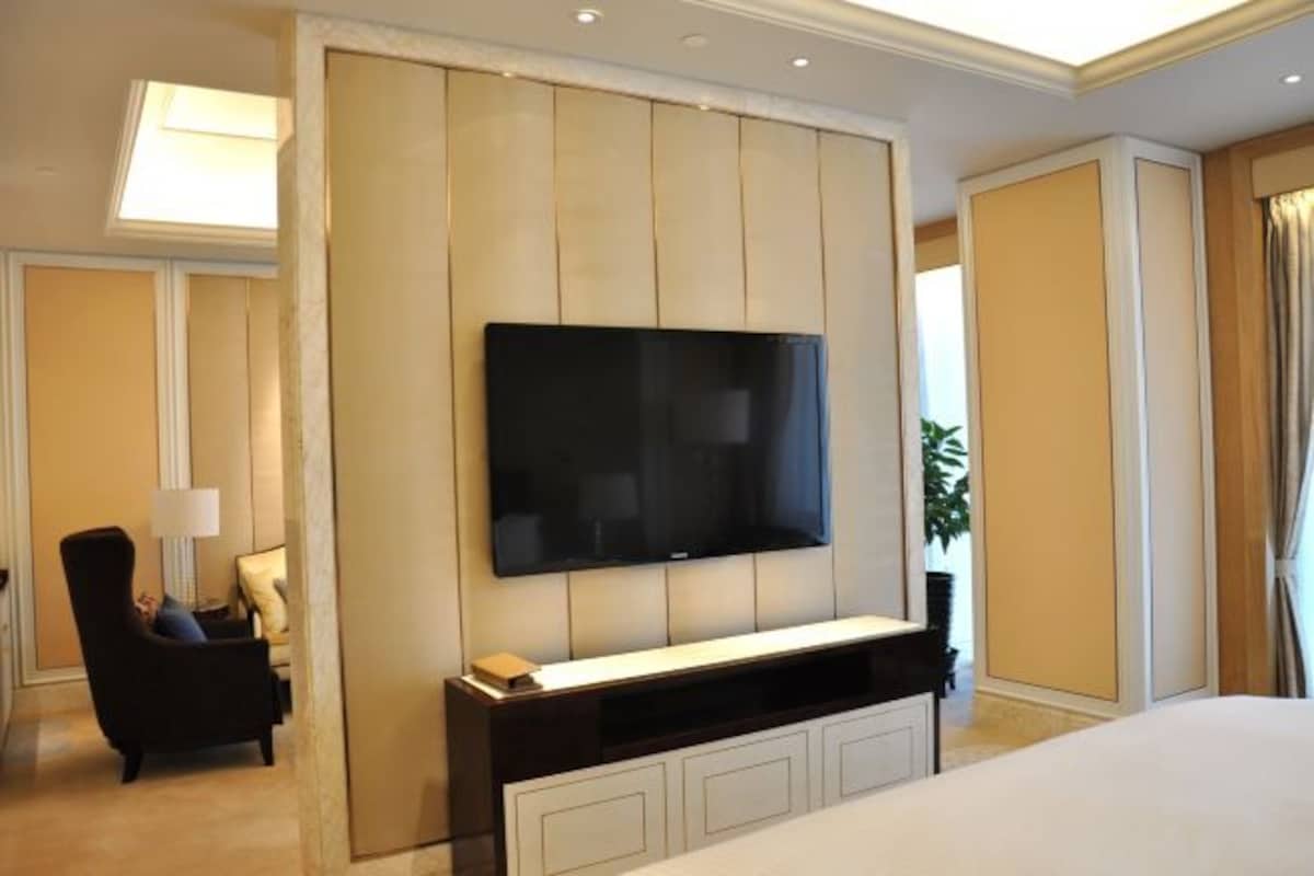 A hotel room with a large flat-screen TV mounted on a wall, a comfortable seating area with a chair and a side table, and a large bed with a white headboard.