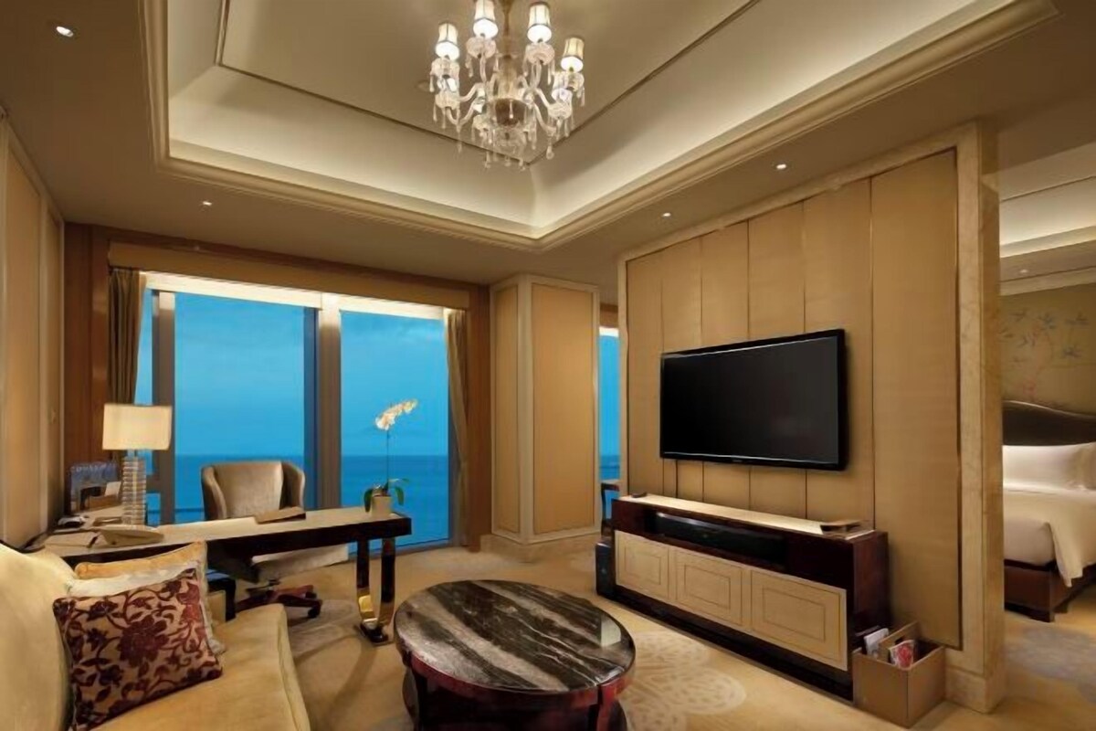 A hotel room with a large flat-screen TV, a seating area with a sofa and a round table, and a view of the ocean through large windows.