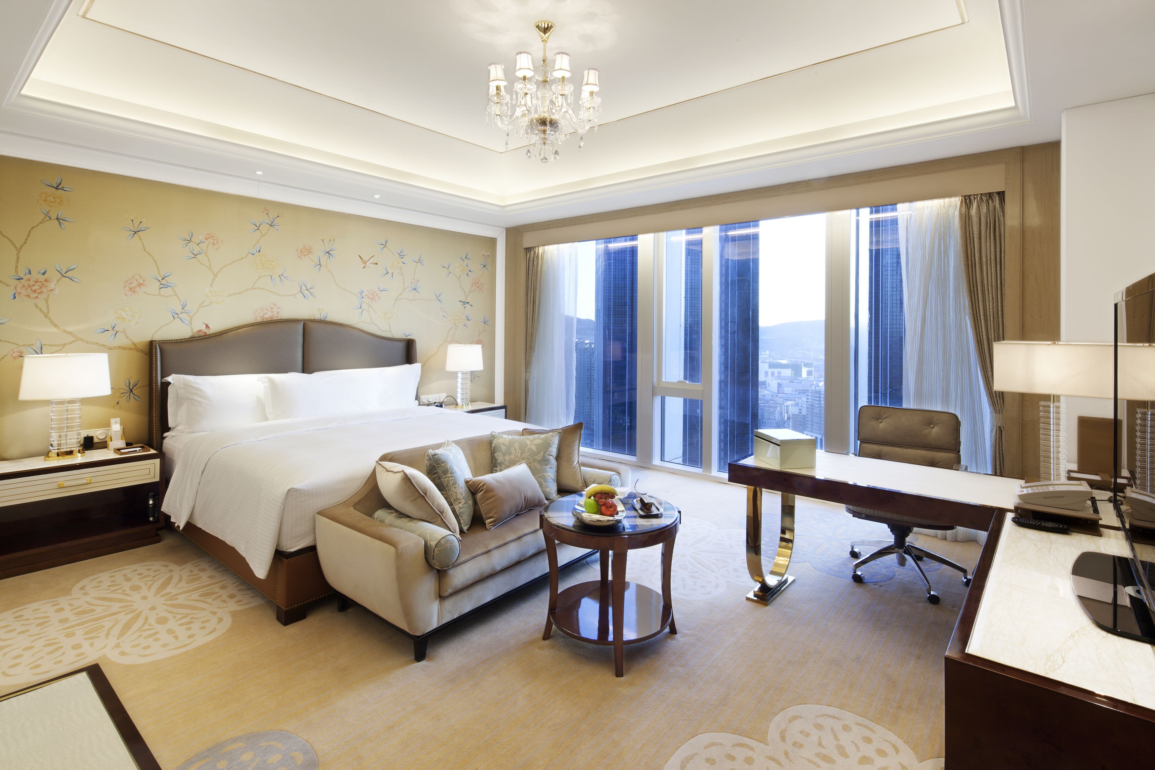 A hotel room with a large bed, a desk, a chair, a sofa, and a view of the city through large windows.