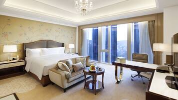 A hotel room with a large bed, a desk, a chair, a sofa, and a view of the city through large windows.