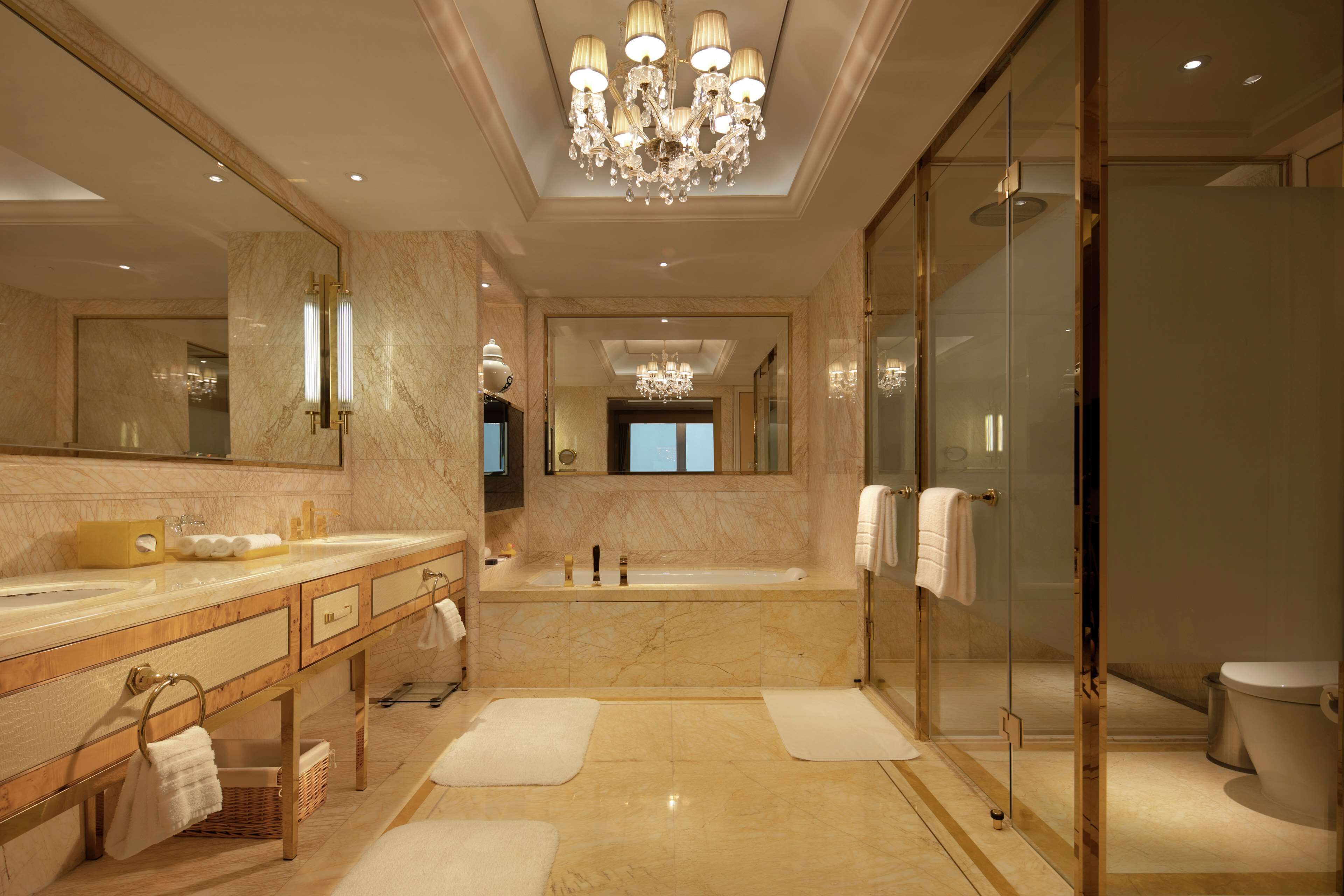 A luxurious bathroom with a marble floor, a large mirror, a chandelier, a bathtub, and a glass-enclosed shower area.