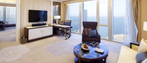 A spacious hotel room with a large flat-screen TV, a comfortable sofa, a dining table, and a balcony with a city view.